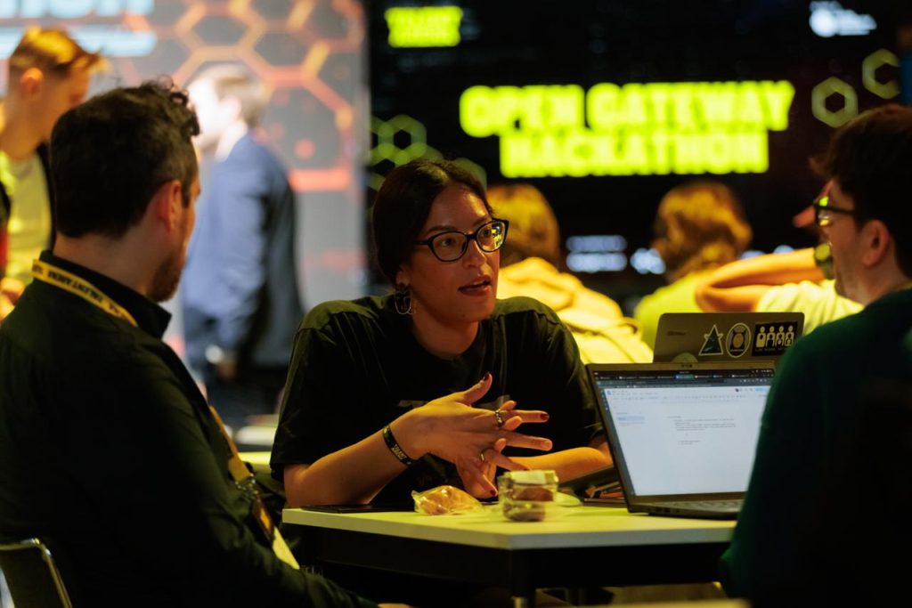 Three people discuss around a table with laptops at an event; "OPEN GATEWAY HACKATHON" on screen behind them.