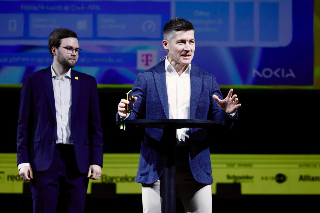 Two men in suits stand on stage; one speaks at a podium holding a trophy, event logos visible behind them.