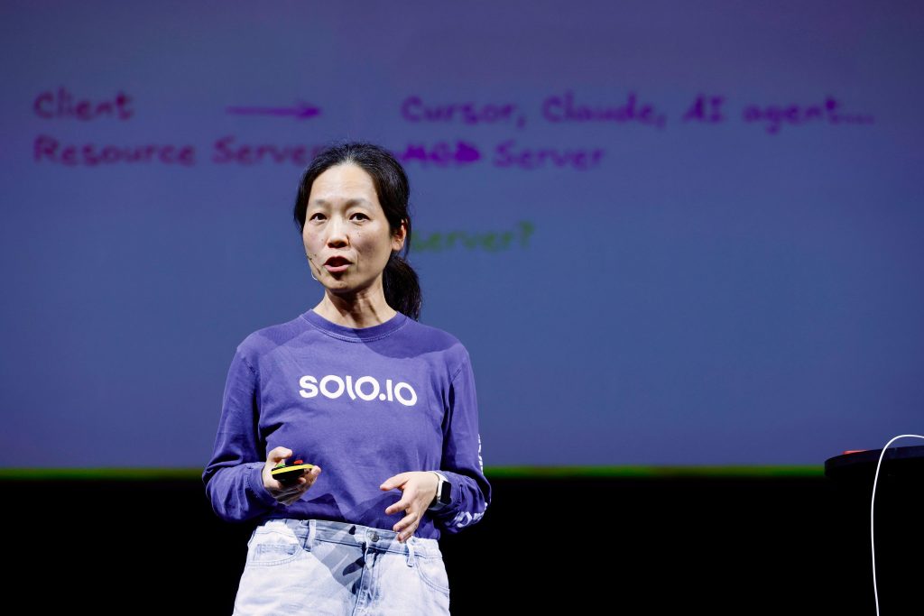 Woman in a purple "solo.io" shirt speaks on stage with a tech-related slide projected behind her.