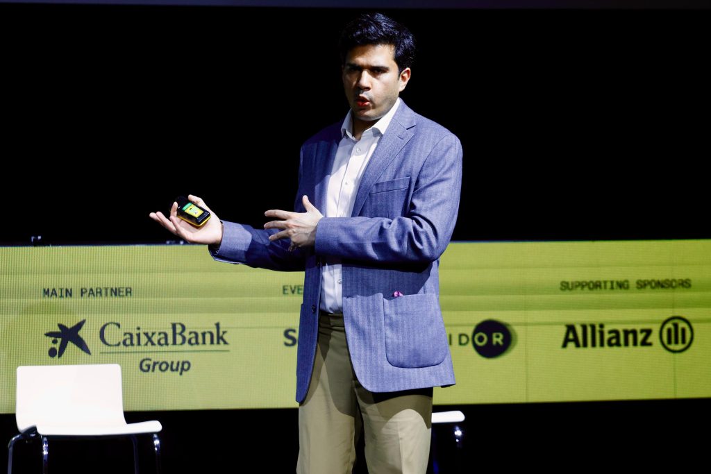 A man in a blazer giving a presentation on stage, gesturing with one hand and holding a device in the other.