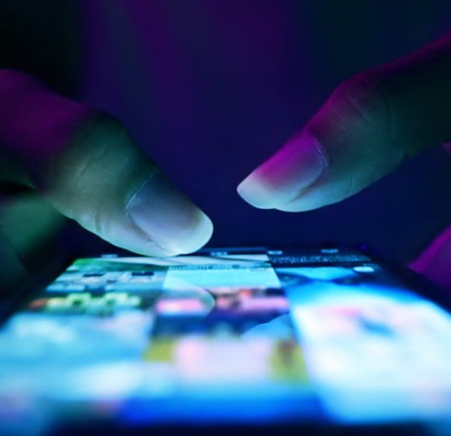 Close-up of hands scrolling on a smartphone screen, illuminated by blue and purple light, capturing the sleek interface of the Skydio app.