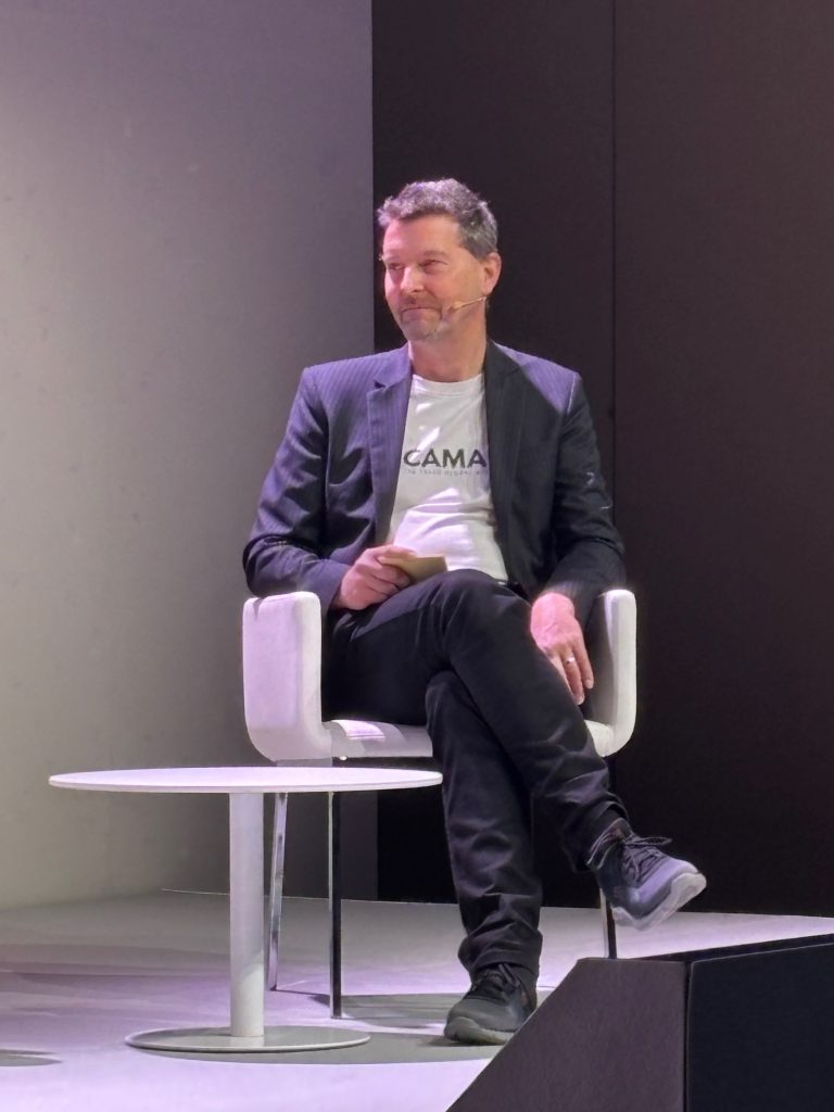 A man in a suit jacket sits on a white chair on stage, holding a notepad, with a small table beside him.
