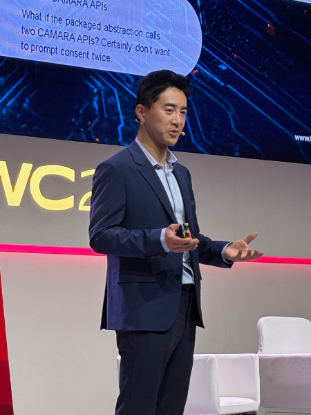 A man in a suit gives a presentation on stage, standing in front of a screen with technical text.