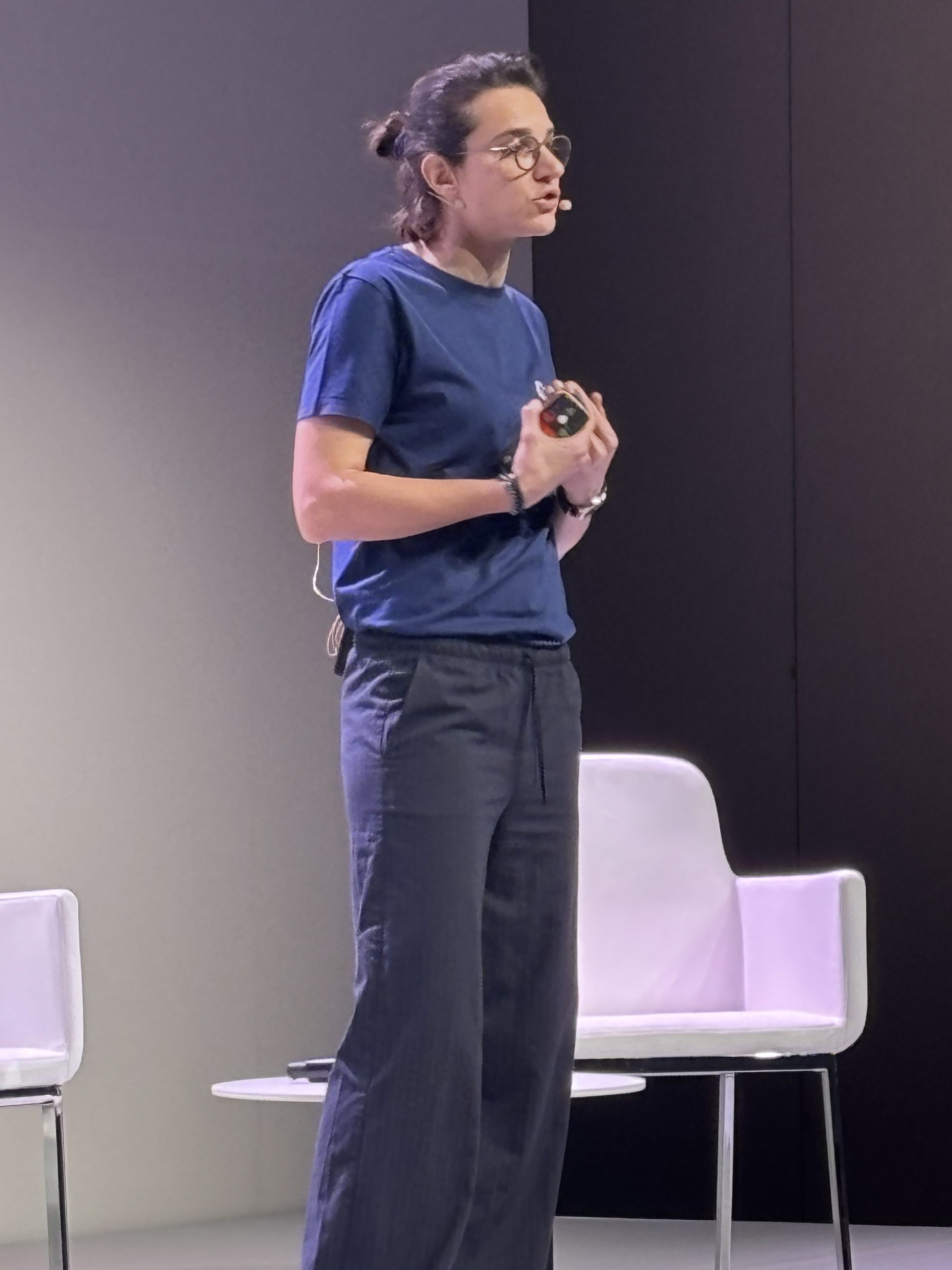 Person in blue shirt and gray pants speaking on stage with a mic, standing next to white chairs.