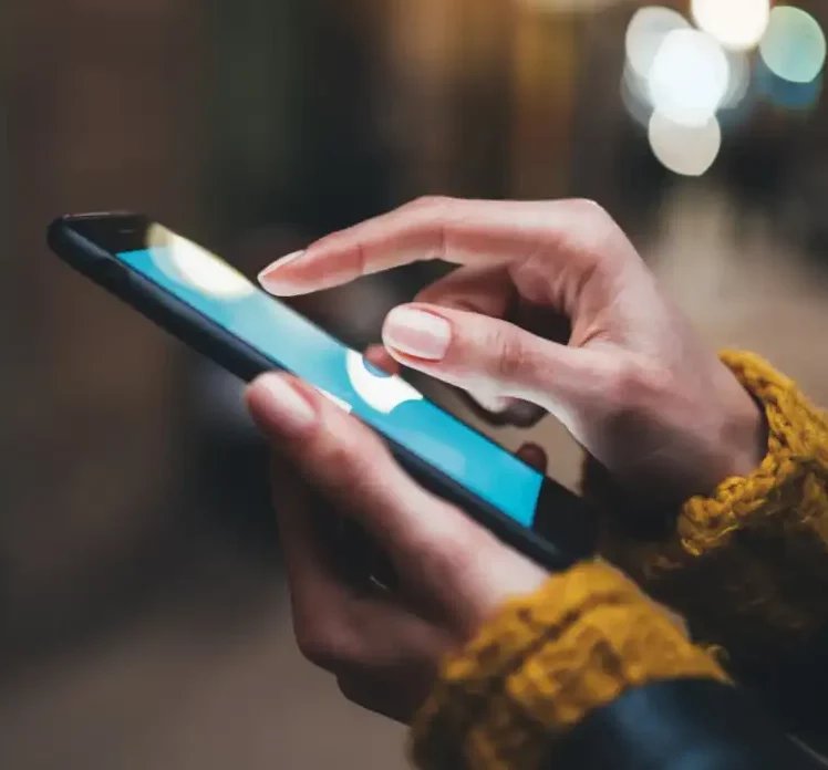 Close-up of hands using a smartphone outdoors at night, with blurred city lights and the reflection of a Skydio drone in the background.