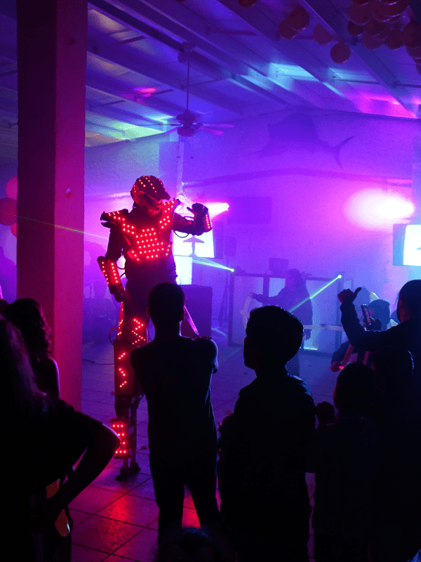 A robot suit covered in red lights performs on a stage with colorful lasers and people watching.