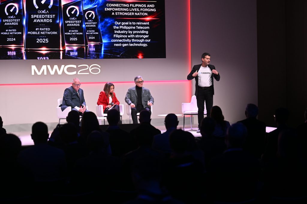 Three people sit on stage while a speaker stands and presents at MWC26, with awards and a message shown on screens.