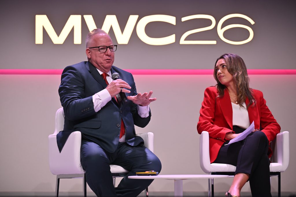 Two people in business attire speak on stage at MWC26, one holding a microphone while the other listens.