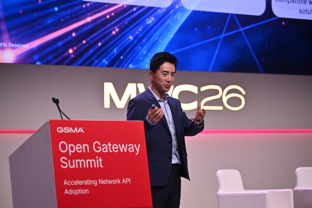 A man presents onstage at the Open Gateway Summit, with "MWC26" and a digital backdrop behind him.