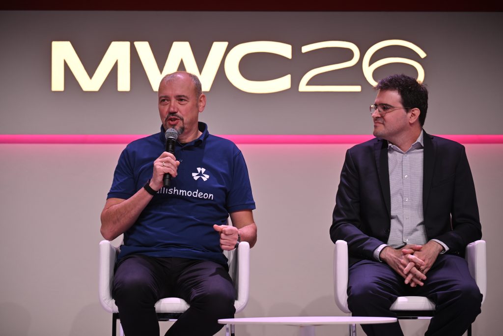 Two men sit on stage; one speaks into a microphone under an "MWC26" sign.