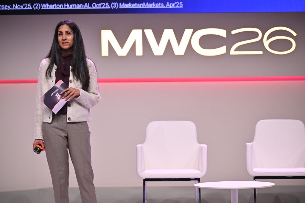 A woman stands on stage at MWC26, holding notes, with two empty chairs behind her—ready to discuss 5G operationalisation and api network monetisation.