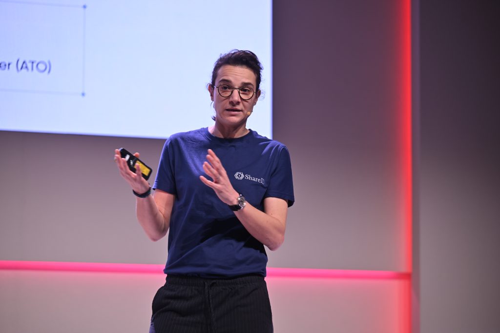 Person wearing glasses and a blue shirt speaks on stage about 5G operationalisation, gesturing with one hand while holding a device.