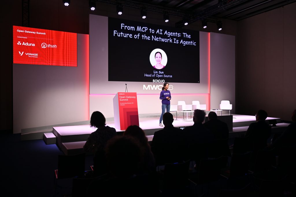 A speaker presents on stage at a tech summit, with a screen displaying a talk on AI agents, 5G operationalisation, and networking.