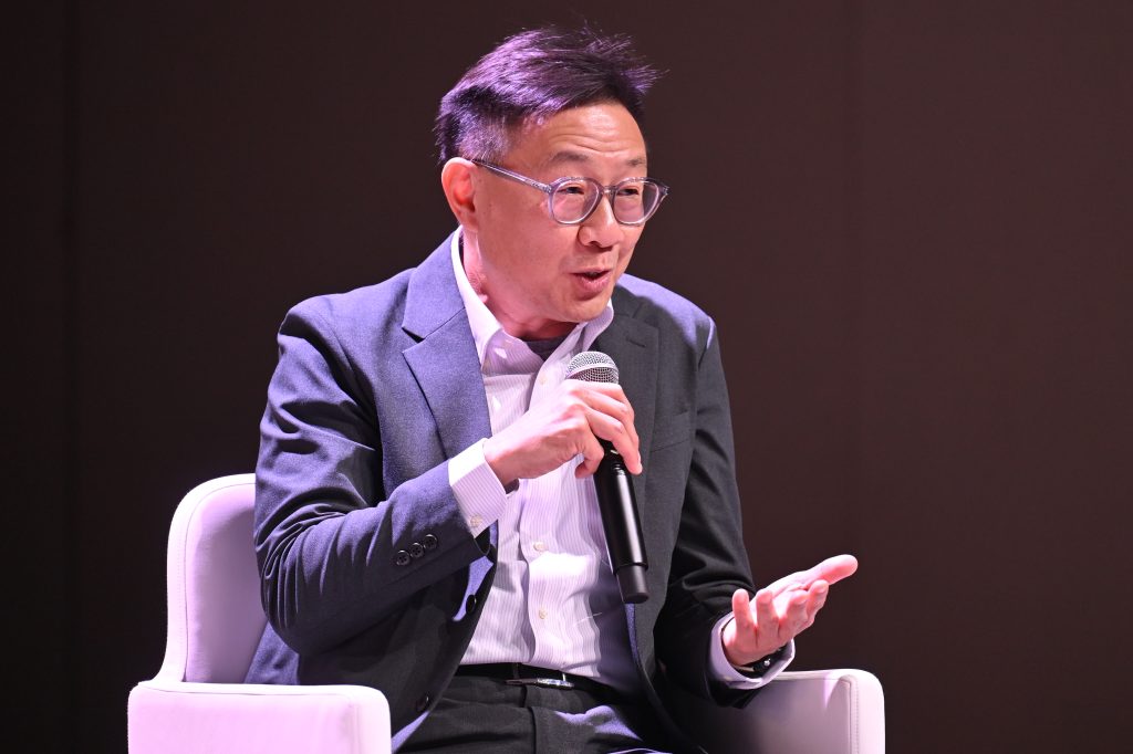 A man in a suit speaks into a microphone while seated on a white chair against a dark background, discussing API network monetisation and 5G operationalisation.