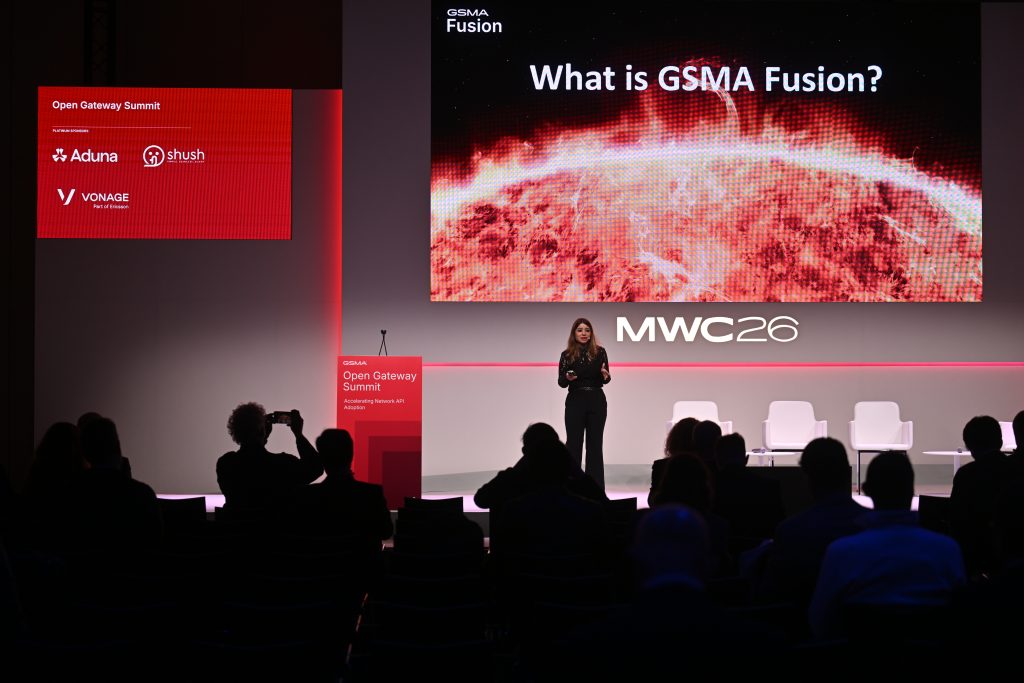 A speaker presents "What is GSMA Fusion?" on stage at MWC26, highlighting API network monetisation, with the audience seated in a dark room.