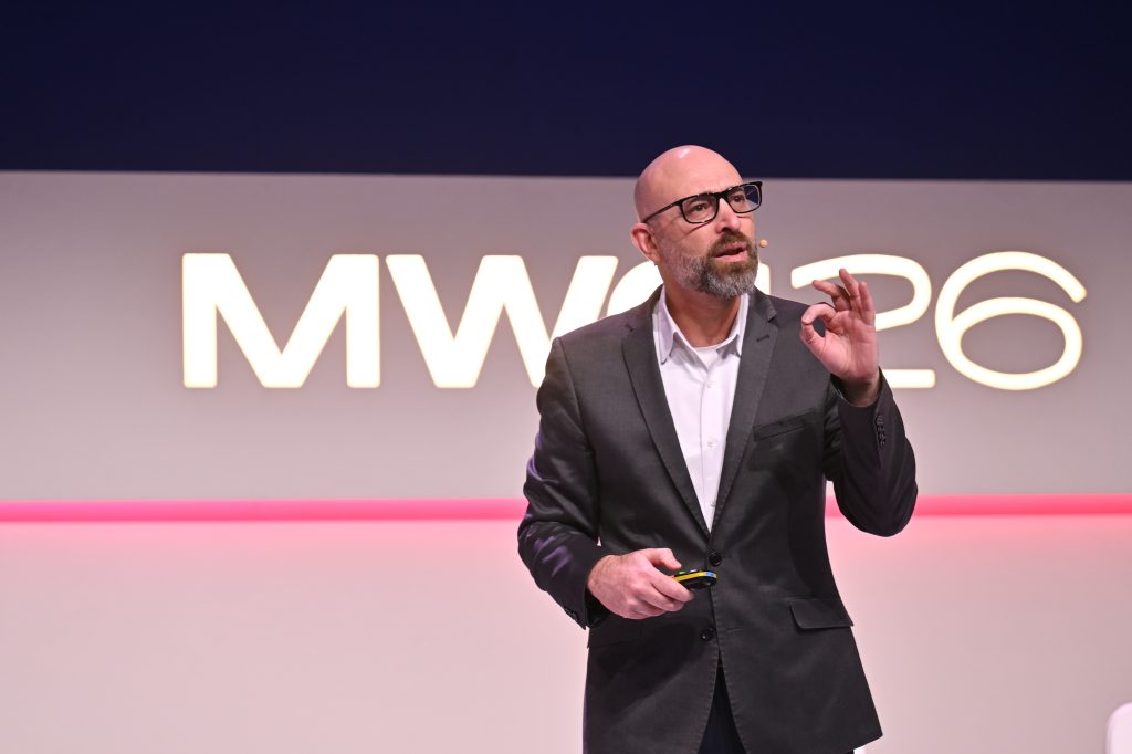 Bald man in a suit speaking on stage in front of a sign reading "MWC 26," discussing 5G operationalisation and api network monetisation.