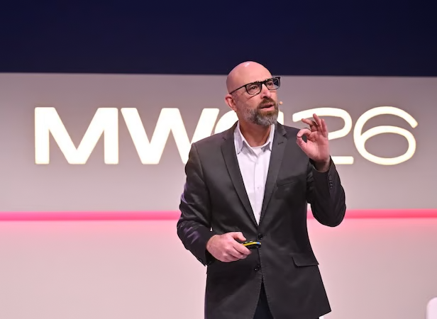 A man in a suit speaks on stage in front of a sign reading "MWC 26," highlighting advancements in 5G operationalisation and api network monetisation.