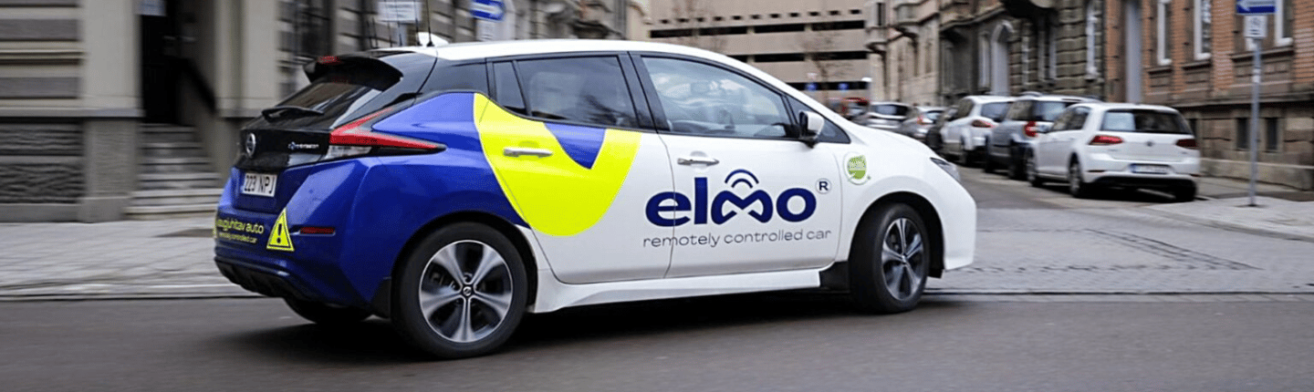 A white and blue Elmo GSMA Fusion remotely controlled car drives on a city street lined with parked cars and buildings.