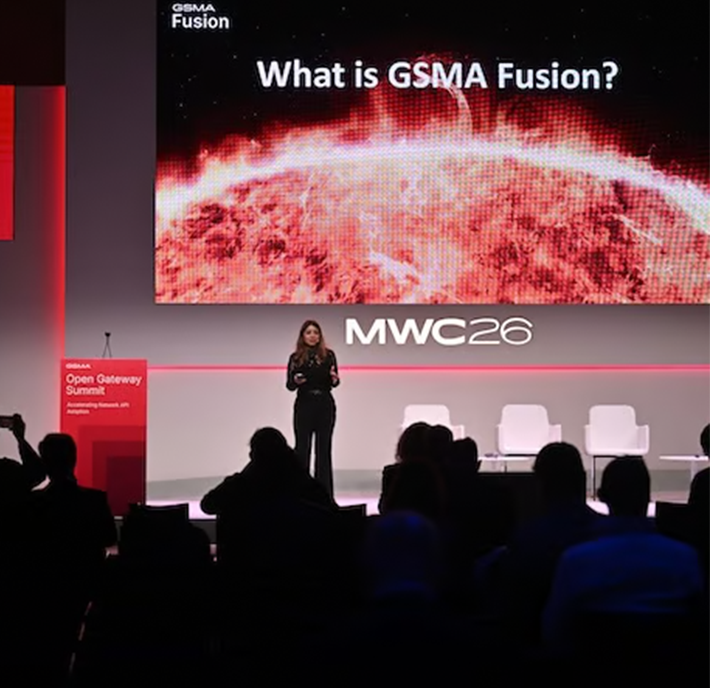 A speaker stands on stage at MWC26 in front of a screen displaying "What is GSMA Fusion?", highlighting how 5G operationalisation and API network monetisation are shaping the future of mobile connectivity.