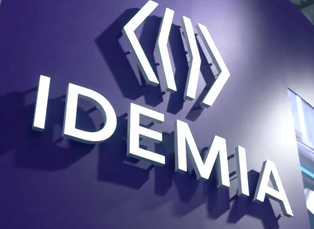 IDEMIA logo in white letters on a purple wall, with geometric shapes above the text.