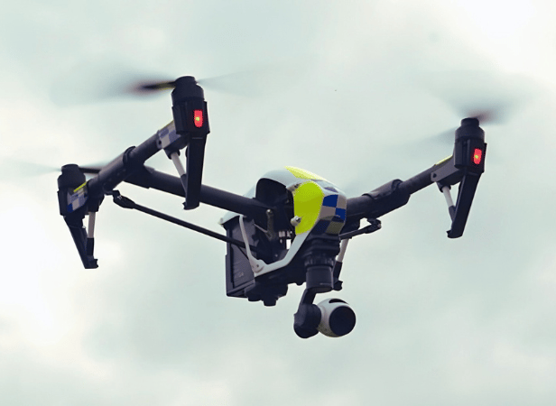 A police drone with a camera hovers in the sky, lights glowing on its arms.