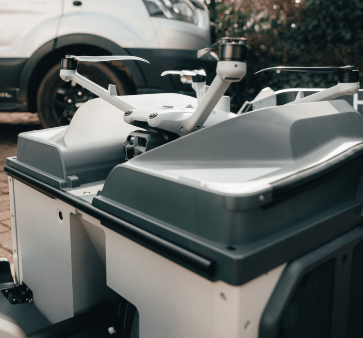 A white drone resting on a gray docking station outdoors, with a vehicle in the background.