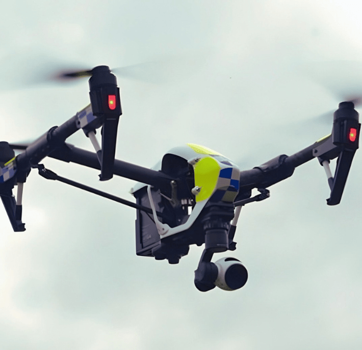A police drone, meeting the statement of requirements, hovers in the cloudy sky with a camera attached. Its yellow and blue checkered pattern, black arms, and red landing lights signal active surveillance or monitoring duties.
