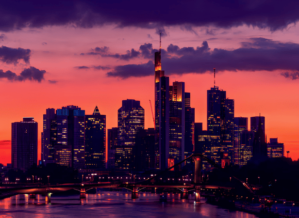 A city skyline at dusk, with tall modern skyscrapers silhouetted against a vibrant orange and purple sunset sky. A river with bridges runs in the foreground, and the city lights begin to illuminate the buildings and water.