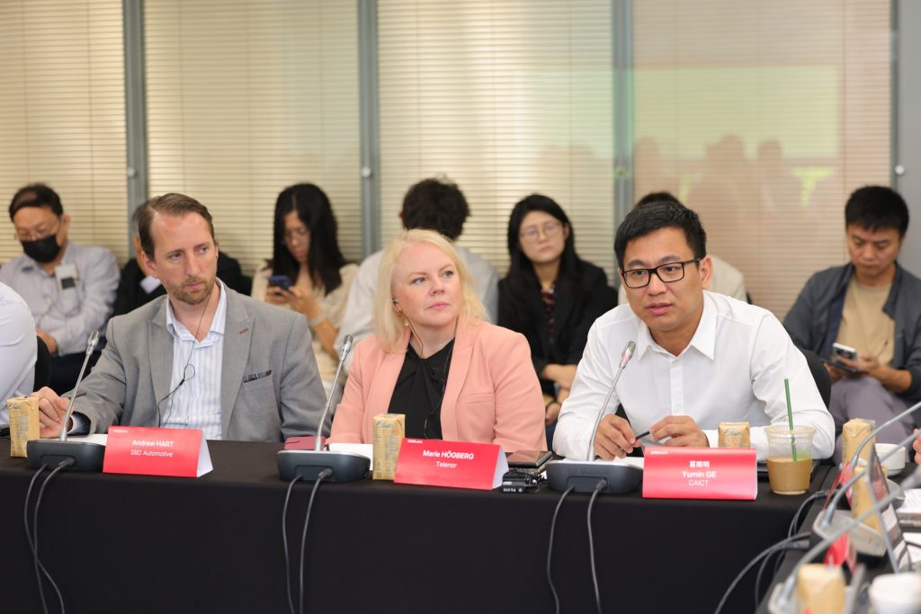 Three people sit at a conference table with microphones, speaking and listening. Others sit behind them, some using phones. Nameplates, drinks, and notepads are on the table. Blinds cover windows in the background. The atmosphere appears formal and focused.
