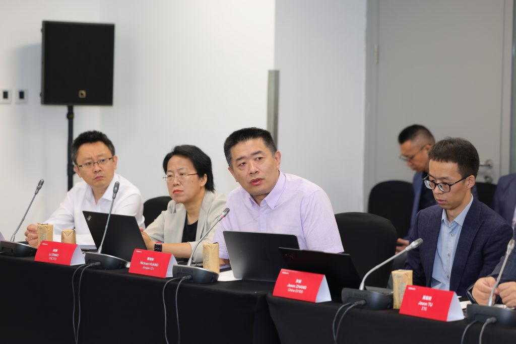 Four people sit at a conference table with laptops and microphones, listening intently. Three face forward, one looks down. Red nameplates are on the table. A speaker stands in the background near a door. The room appears modern and professional.