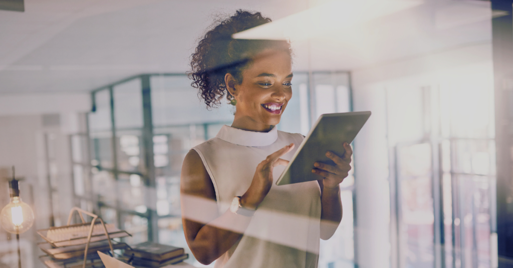A woman stands in a modern office, smiling while using a tablet to explore Network APIs. She wears a sleeveless white top and has curly hair pulled back. Sunlight filters through glass walls, with books and office supplies visible in the background.