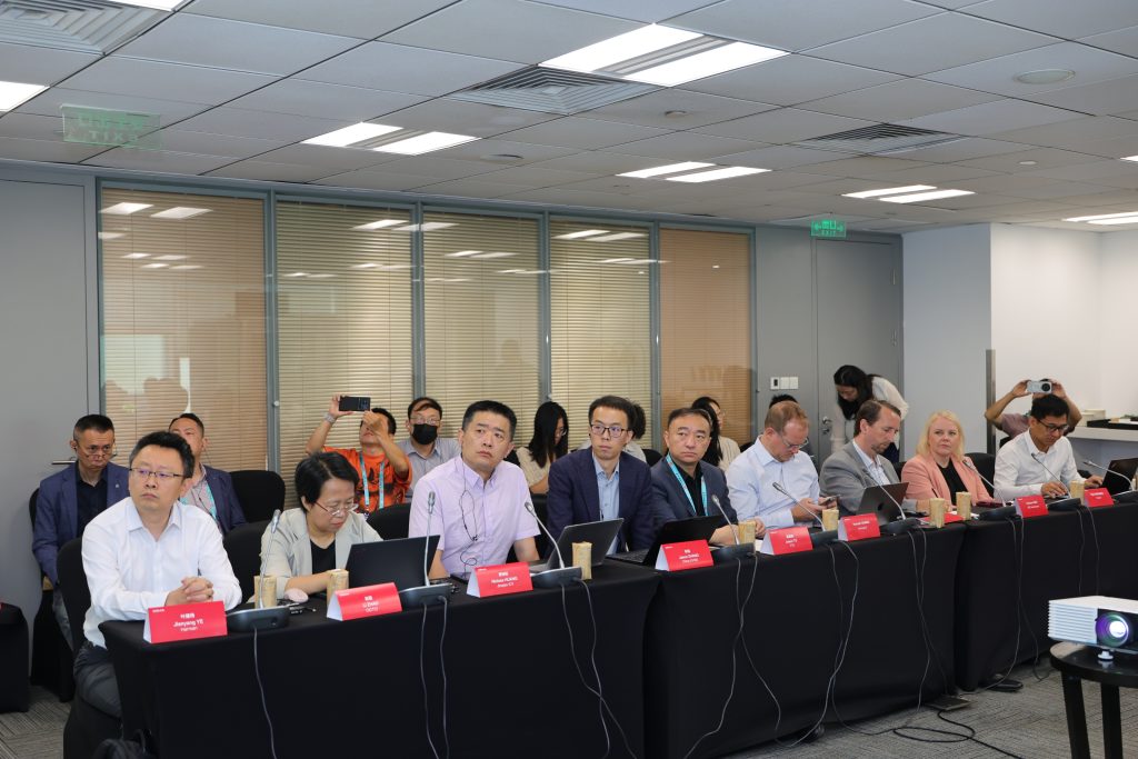A group of people sit at a long table in a modern conference room, facing forward at the MWC Shanghai 2026 - ICV Automotive Roundtable. Each person has a laptop and a red nameplate. Some participants listen attentively, while others take notes or photos. The background reveals glass walls and additional office space, emphasizing the event’s professional atmosphere.