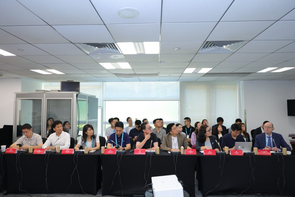 A diverse group of people sit at two long black tables with red nameplates, facing forward in a modern conference room with white walls, large windows, and ceiling lights. Some participants use laptops; others listen or watch attentively during the MWC Shanghai 2026 - ICV Automotive Roundtable.