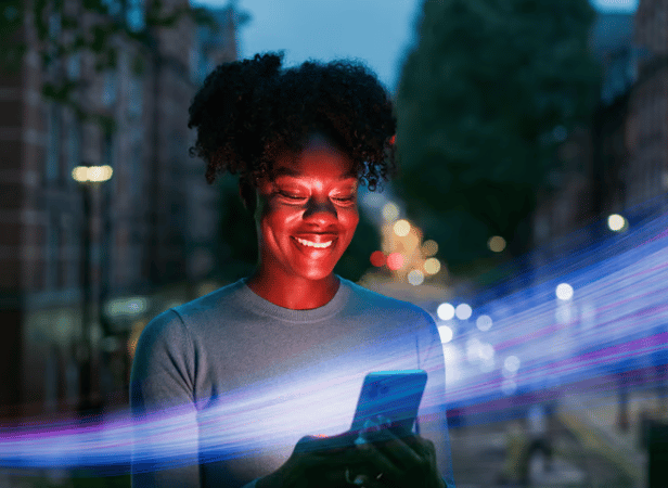 A smiling woman stands outdoors at dusk, looking at her smartphone. City lights and blurred car headlights are visible in the background. Blue and pink light streaks cross the image, suggesting technology or digital connection.