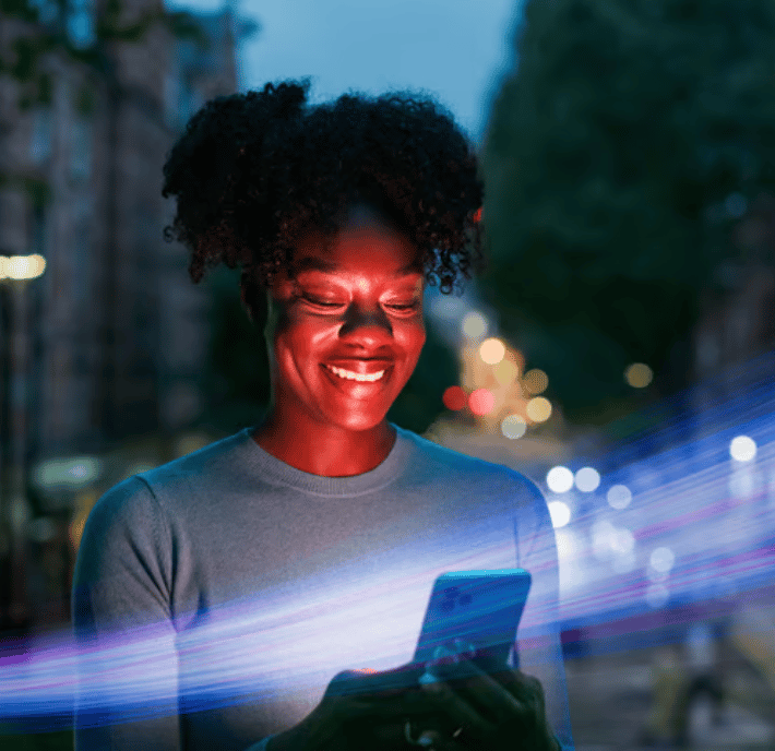 A smiling woman stands outdoors at dusk, looking at her phone as she reviews a statement of requirements. City lights blur in the background. Blue and purple light streaks cross the foreground, illuminating her face with a soft glow and creating a futuristic, vibrant atmosphere.