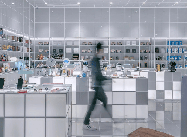 A person in motion walks through a futuristic electronics store with white, illuminated display tables and shelves showcasing gadgets, tech devices, and boxed products. The space is bright, modern, and tidy, with reflective white tiles on the floor and walls.