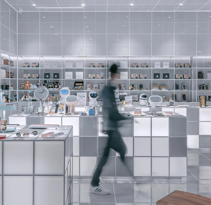 A person in motion walks through a modern, minimalistic store with white and gray tiles. Shelves behind the counter display electronics, toys, and gadgets that meet every statement of requirements for quality and innovation. The blurred figure wears a dark outfit and white sneakers. Counters display small tech items.