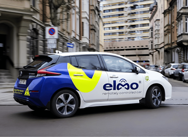 A blue and white car labeled “elmo remotely controlled car” drives on a city street lined with buildings. The vehicle features yellow graphics and a caution triangle on the rear, indicating it’s a remote-controlled or self-driving vehicle.