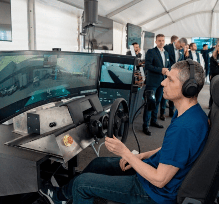 A man in headphones sits at a driving simulator with three large monitors displaying a road scene. He holds a steering wheel and wears a blue shirt. Several people in business attire stand in the background inside a white tent.