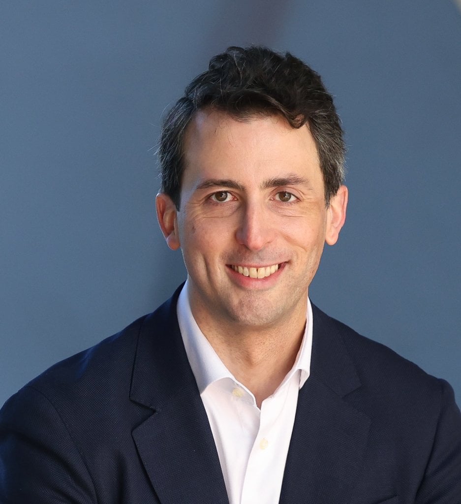 Smiling man with short dark hair, wearing a white shirt and dark blazer, against a plain blue-gray background.