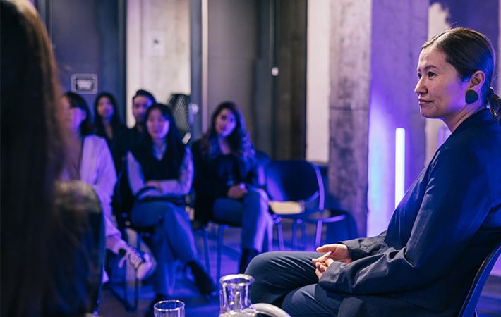 In a dimly lit room with a modern aesthetic, a woman in a blue outfit discusses mobile fintech before an attentive audience. Mostly women, the audience sits in chairs facing her as cool, ambient blue lighting highlights the space.