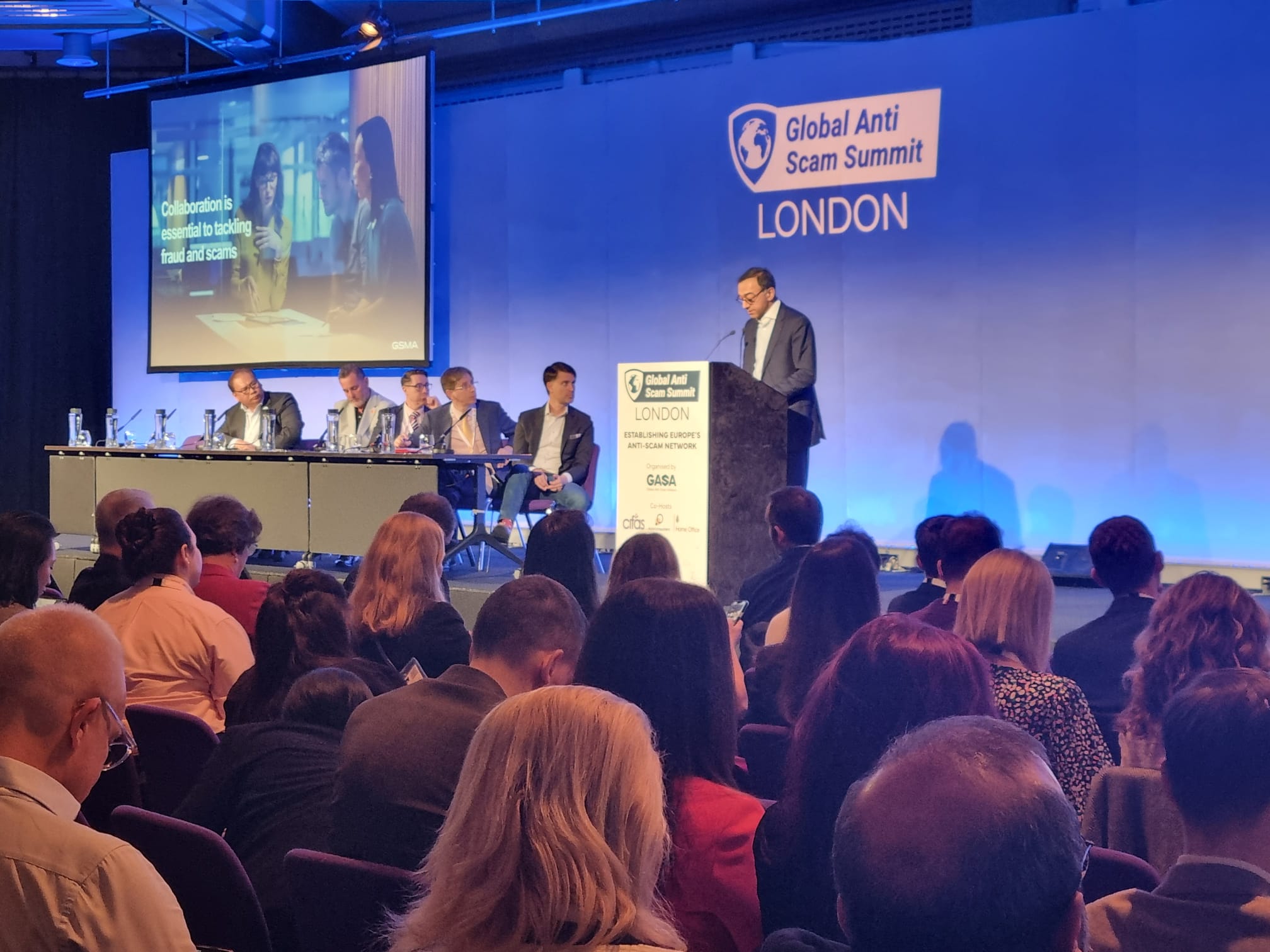 A speaker stands at a podium on a stage at the Global Anti Scam Summit in London, where mobile fintech solutions are being discussed. Behind him, a large screen displays his presentation. Seated to the left are five panelists in suits, while the engaged audience is visible in the foreground.