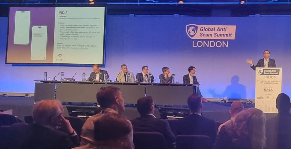 A speaker at the Global Anti Scam Summit in London presents a slide labeled "INDIA" on a large screen. Five panelists sit at a long table on stage. Attendees face forward, focusing on the presentation. The room is dimly lit with blue hues.