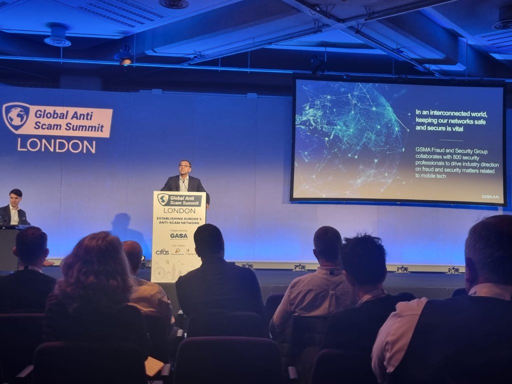 A speaker stands at a podium on a stage delivering a presentation at the Global Anti Scam Summit in London. A large screen displays a slide that discusses the importance of network security. The audience is seated, facing the stage. The room is dimly lit with blue lighting.