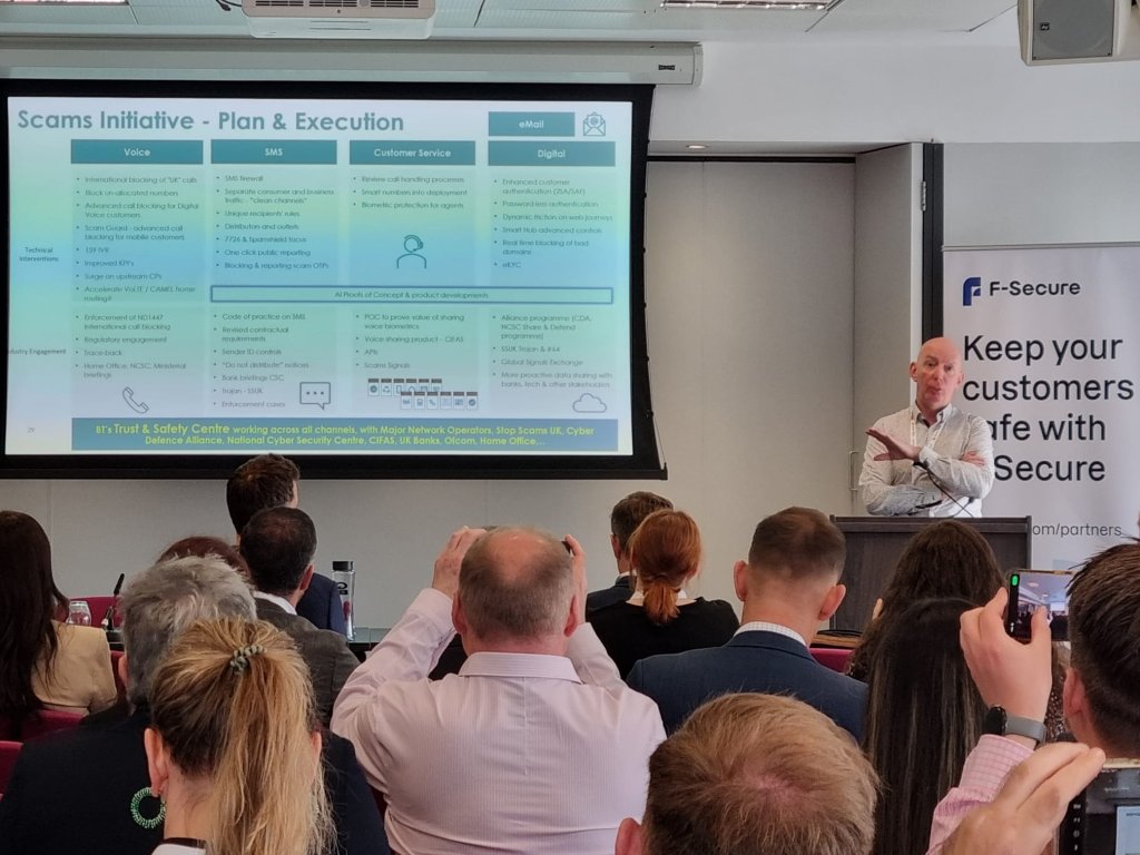 A man stands at a podium giving a presentation in a conference room. Behind him is a large slide titled "Scams Initiative - Plan & Execution." Attendees are seated, some taking photos with their phones. A banner with "F-Secure" and text about customer safety is on the right wall.