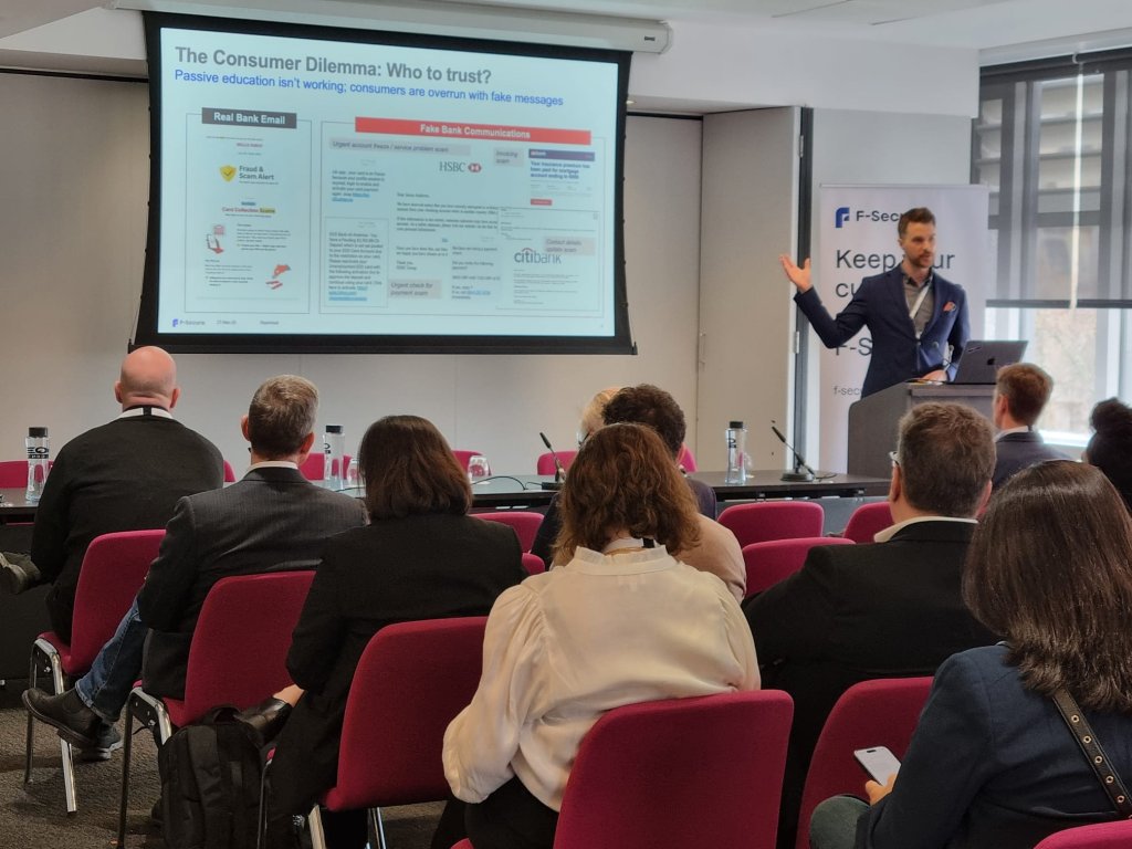 A man in a suit presents a slide labeled "The Consumer Dilemma: Who to trust?" to an audience in a conference room. The audience sits in rows of red chairs, looking at the screen. A banner with "F-Secure" is visible on the right. The room has a modern and professional atmosphere.