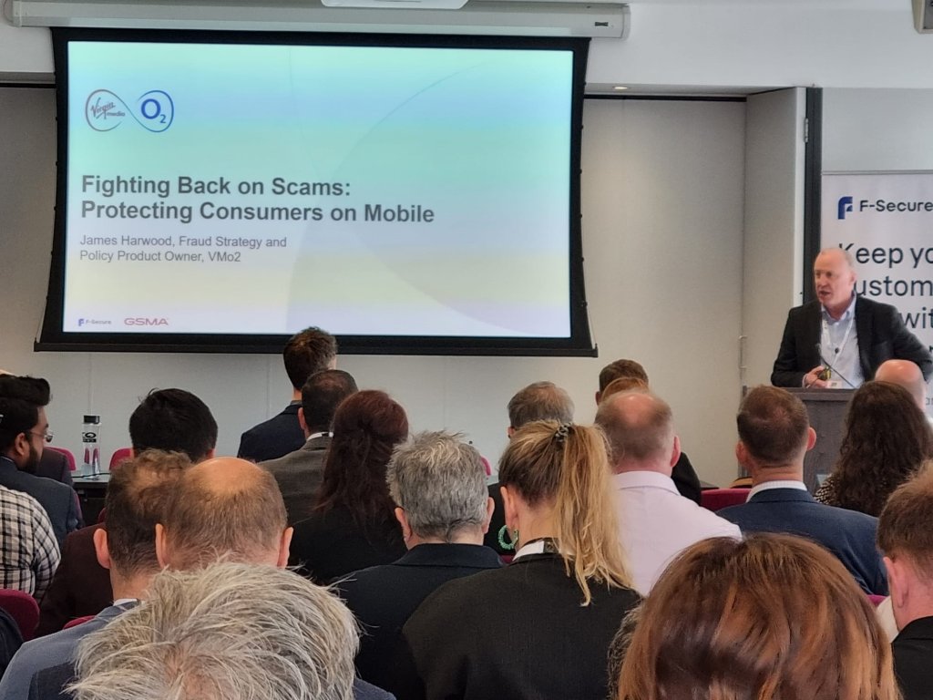 A speaker addresses an audience at a conference. A slide titled "Fighting Back on Scams: Protecting Consumers on Mobile" is displayed, with logos of Virgin and O2. Attendees sit facing the screen, and the room has a professional setting with branding visible on the walls.
