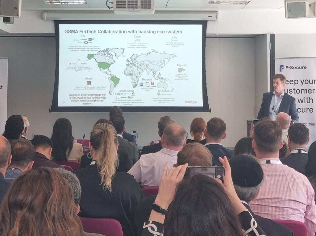 A speaker presents at a conference in a room filled with people. A slide titled "GSMA FinTech Collaboration with banking eco-system" displays a world map, highlighting areas in the US, India, Nigeria, and South Africa. Attendees are seated, and one person in the foreground takes a photo.