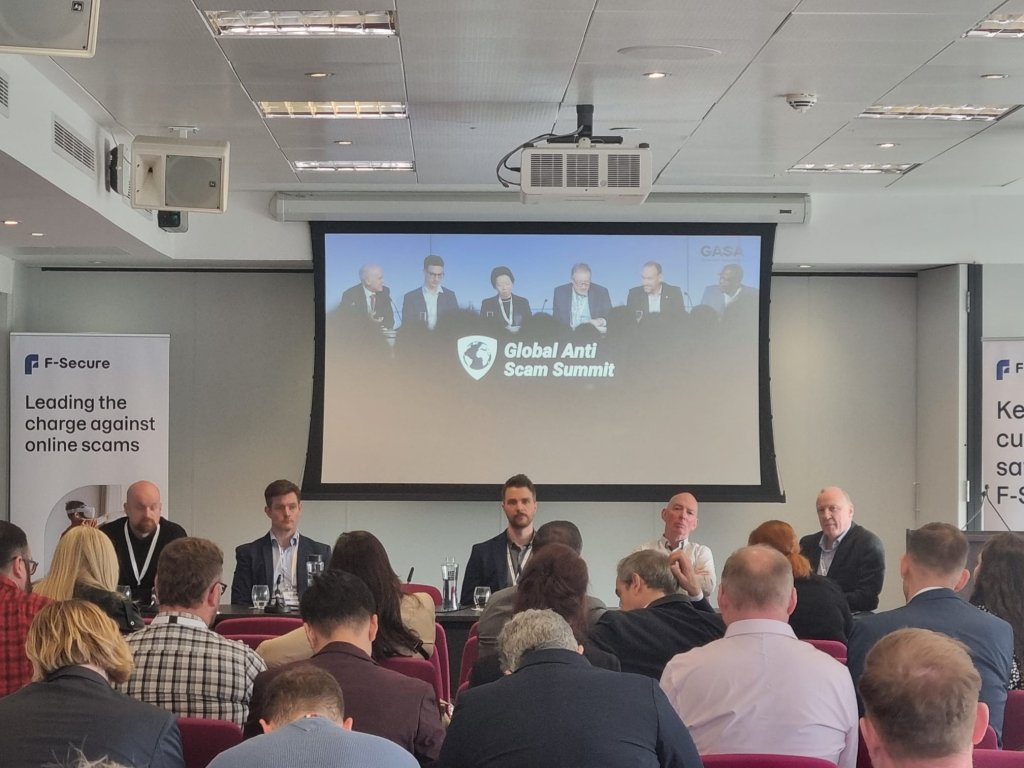 A conference room with six panelists on stage, seated behind a table. The screen above displays "Global Anti Scam Summit." Attendees are seated, facing the panel. Banners read "F-Secure: Leading the charge against online scams." The room is well-lit and modern.