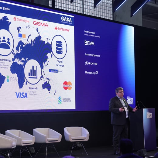 A man in a suit presents on stage at a fintech forum, beside a large screen displaying a world map, company logos (Visa, Mastercard, Santander), and business charts. Three empty white chairs and sponsor banners like BBVA and Chainalysis are visible.